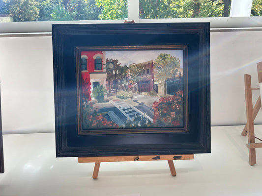 Framed painting on an easel with a scenic view of buildings and trees.