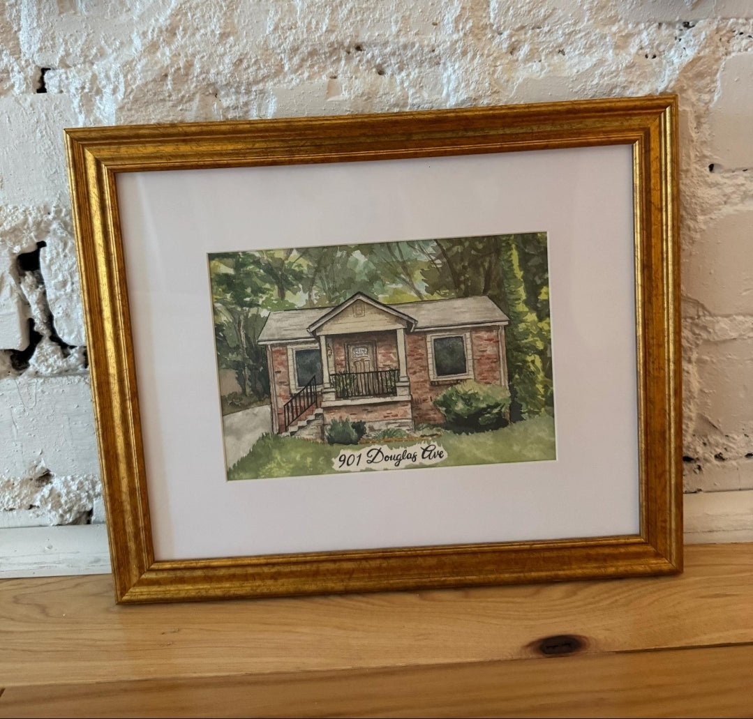 A framed custom watercolor painting of a brick house with a porch, matted in white and enclosed in a gold frame, resting on a wooden surface against a white brick wall.