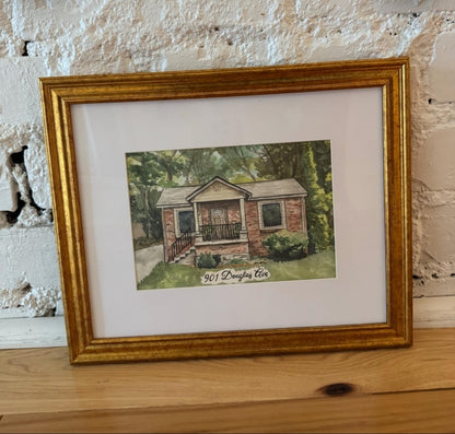 A framed custom watercolor painting of a brick house with a porch, matted in white and enclosed in a gold frame, resting on a wooden surface against a white brick wall.