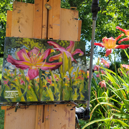A painted flower scene on an easel in a garden. The artwork shows pink and yellow daylilies with green stems and foliage, while real flowers and greenery are visible around it.