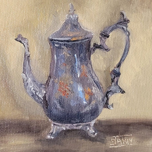A framed still life painting of a blue-gray teapot on a warm beige background, enclosed in an ornate gold and blue frame mounted on a light gray wall.